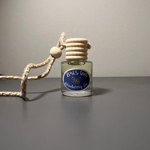 Op de afbeelding: Een heldere glazen fles met een houten dop en een blauw-wit etiket met de tekst "Bui's Tees Blueberry Pie". De fles hangt aan een gouden en witte touwtje.