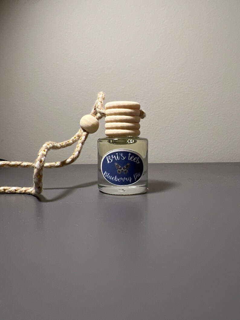 Op de afbeelding: Een heldere glazen fles met een houten dop en een blauw-wit etiket met de tekst "Bui's Tees Blueberry Pie". De fles hangt aan een gouden en witte touwtje.