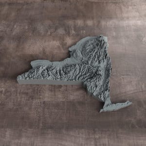 May include: A three-dimensional gray relief map of New York State featuring detailed topographical elevations and valleys. The physical model displays the varied terrain of the region with a matte finish, highlighting the mountain ranges and plains.