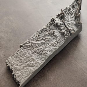 May include: A gray 3D printed topographic map model featuring a detailed relief of a mountain range with deep valleys and jagged peaks. The rectangular object displays intricate terrain textures and elevation changes across its surface.