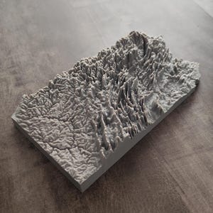 May include: A rectangular gray 3D printed topographic map model featuring intricate raised terrain details, including mountain ridges, valleys, and river systems, set on a flat base with a textured, matte surface finish.