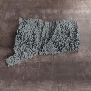May include: A gray, three-dimensional topographic map of the state of Connecticut. The model features a highly detailed, textured surface representing the varied elevation and terrain of the region, cast in a solid, monochromatic gray finish.