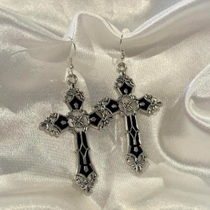 May include: A pair of silver-toned cross earrings with black enamel detailing. The crosses have an ornate, gothic design with decorative flourishes. The earrings hang from simple silver hooks, set against a white, silky background.