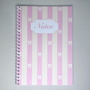 May include: A spiral-bound notebook with a pink and white striped pattern. The cover features a decorative label with the word "Notes" in script. Small bow illustrations are evenly spaced across the cover.