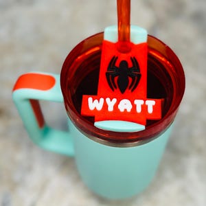 May include: A teal insulated tumbler with a red lid and handle accents. The tumbler features a red and black Spider-Man-themed charm with the name "WYATT" in white letters. A clear orange straw is inserted into the lid.