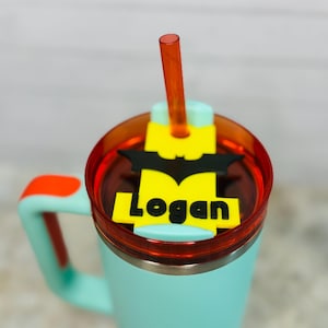 May include: A teal insulated tumbler with a red lid and handle accents. The lid features a yellow and black Batman logo with the name "Logan" in black letters. An orange straw extends from the lid.