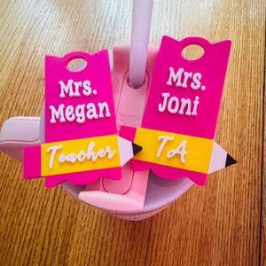 May include: Two pink pencil-shaped drink charms with white text that reads "Mrs. Megan Teacher" and "Mrs. Joni TA". The charms have a yellow band and a pencil tip design. The charms are attached to a pink cup with a straw.