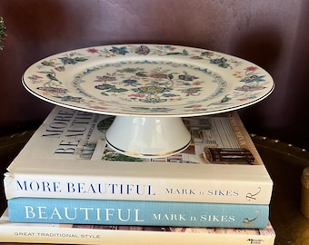 May include: A white porcelain cake stand with a floral patterned plate. The plate has a gold rim and is supported by a white pedestal base. The stand rests on a stack of books with the titles "MORE BEAUTIFUL" and "BEAUTIFUL".