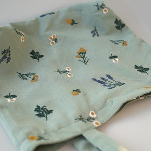 May include: A light green bonnet with a floral pattern. The fabric is decorated with small illustrations of yellow, white, and purple flowers, and green leaves. The bonnet has ties on each side.