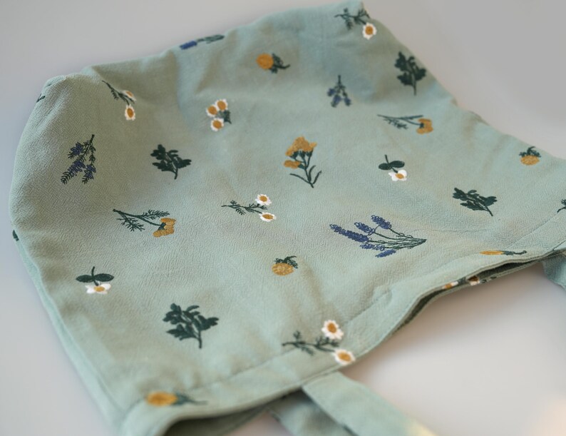 May include: A light green bonnet with a floral pattern. The fabric is decorated with small illustrations of yellow, white, and purple flowers, and green leaves. The bonnet has ties on each side.