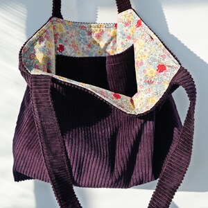 Op de afbeelding: Een dieppaarse corduroy tote bag met een bloemenpatroon voering. De tas heeft een rechthoekige vorm met een brede opening en lange bandjes. De binnenvoering heeft een kleurrijke bloemenprint met rode, gele en blauwe bloemen.