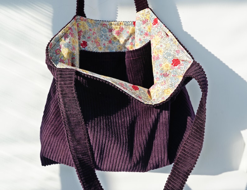Op de afbeelding: Een dieppaarse corduroy tote bag met een bloemenpatroon voering. De tas heeft een rechthoekige vorm met een brede opening en lange bandjes. De binnenvoering heeft een kleurrijke bloemenprint met rode, gele en blauwe bloemen.