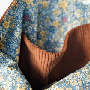 May include: Close-up of a brown corduroy tote bag interior with a floral patterned lining. The lining features a blue background with small yellow and white flowers. The bag has a pocket and a textured corduroy exterior.