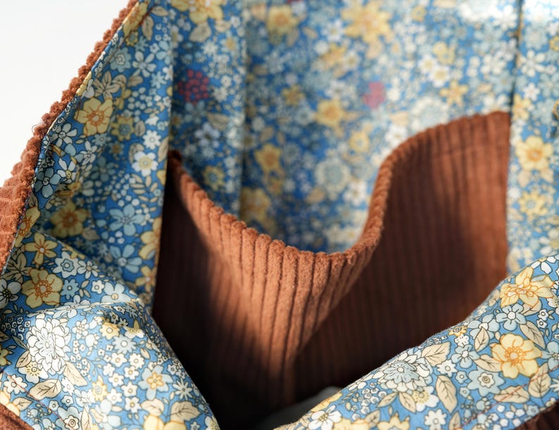 May include: Close-up of a brown corduroy tote bag interior with a floral patterned lining. The lining features a blue background with small yellow and white flowers. The bag has a pocket and a textured corduroy exterior.