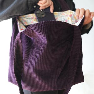 Op de afbeelding: Een dieppaarse corduroy tote bag met lange banden. De binnenkant van de tas heeft een bloemenpatroon voering. Een persoon plaatst een donkergekleurde telefoon in de zak van de tas.