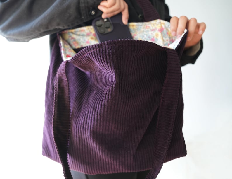 Op de afbeelding: Een dieppaarse corduroy tote bag met lange banden. De binnenkant van de tas heeft een bloemenpatroon voering. Een persoon plaatst een donkergekleurde telefoon in de zak van de tas.