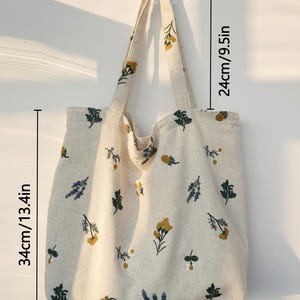 May include: A beige tote bag with floral embroidery. The bag is 13.4 inches wide, 13.4 inches tall, and has a strap that is 9.5 inches long. The bag features yellow, green, and blue floral designs.