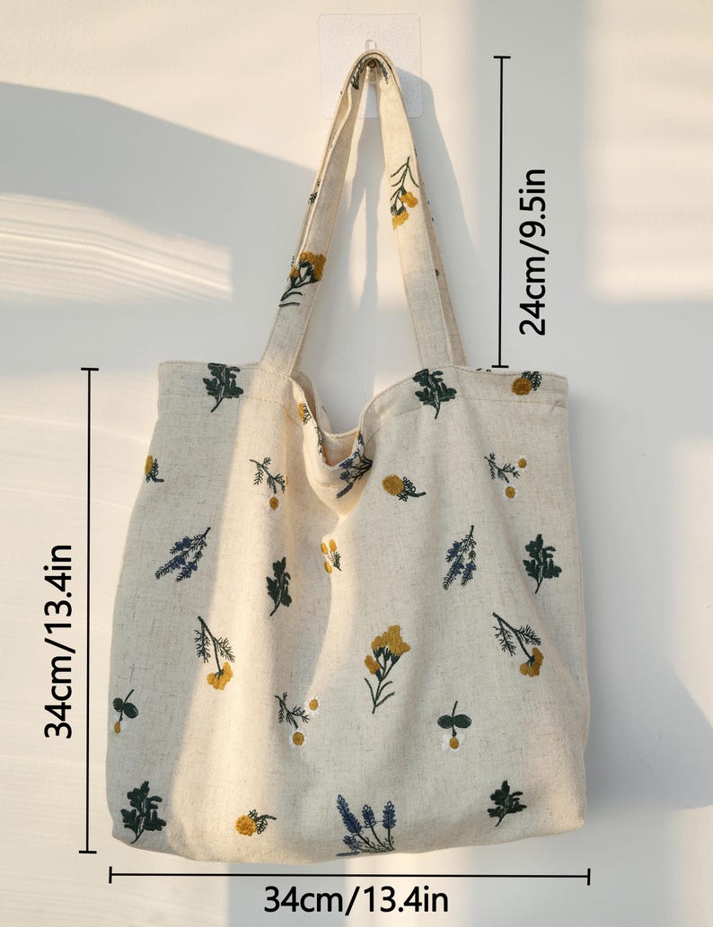 May include: A beige tote bag with floral embroidery. The bag is 13.4 inches wide, 13.4 inches tall, and has a strap that is 9.5 inches long. The bag features yellow, green, and blue floral designs.