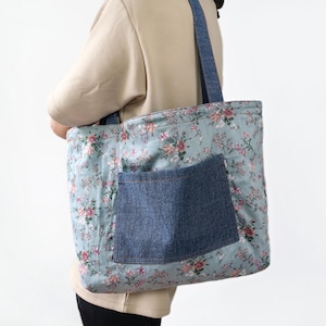 May include: A large tote bag featuring a light blue fabric with a repeating floral pattern of pink and white flowers. The bag includes a dark blue denim exterior pocket and matching dark blue denim shoulder straps for carrying the item.