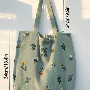 May include: A sage green tote bag with embroidered floral designs. The bag measures 13.4 inches wide, 13.4 inches tall, and has 9.5-inch handles. The floral embroidery includes yellow, white, and purple flowers.