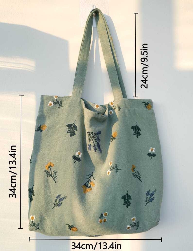 May include: A sage green tote bag with embroidered floral designs. The bag measures 13.4 inches wide, 13.4 inches tall, and has 9.5-inch handles. The floral embroidery includes yellow, white, and purple flowers.