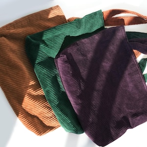 Op de afbeelding: Vier corduroy tote bags in bruin, groen en paars worden getoond. E&eacute;n tas heeft een gestreepte band in groen, wit en bruin. De tassen zijn gemaakt van een geribbelde corduroy stof.