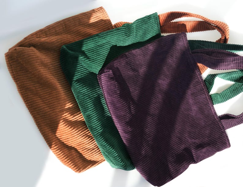 Op de afbeelding: Vier corduroy tote bags in bruin, groen en paars worden getoond. E&eacute;n tas heeft een gestreepte band in groen, wit en bruin. De tassen zijn gemaakt van een geribbelde corduroy stof.