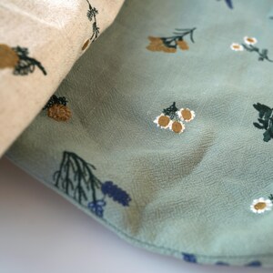 May include: Close-up of two fabrics, one beige and one teal, both with embroidered floral patterns. The beige fabric features brown and green pinecone-like designs. The teal fabric has white, brown, and green floral and leaf patterns.