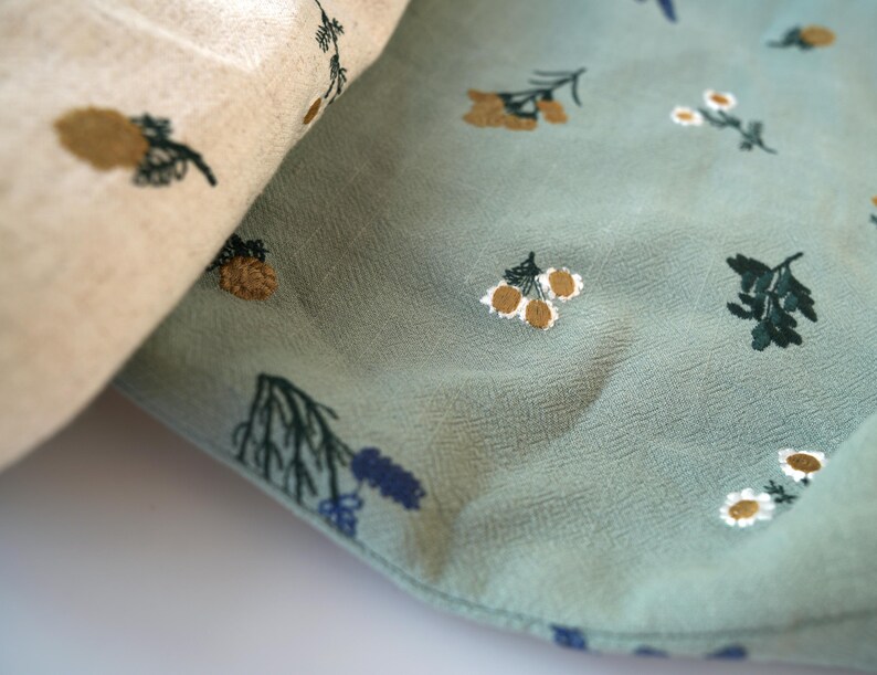 May include: Close-up of two fabrics, one beige and one teal, both with embroidered floral patterns. The beige fabric features brown and green pinecone-like designs. The teal fabric has white, brown, and green floral and leaf patterns.