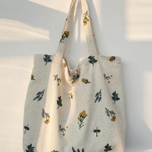 May include: Cream-colored tote bag with embroidered floral patterns in yellow, green, and blue. The bag has two long handles and is made of a textured fabric. The floral design is spread across the bag's surface.