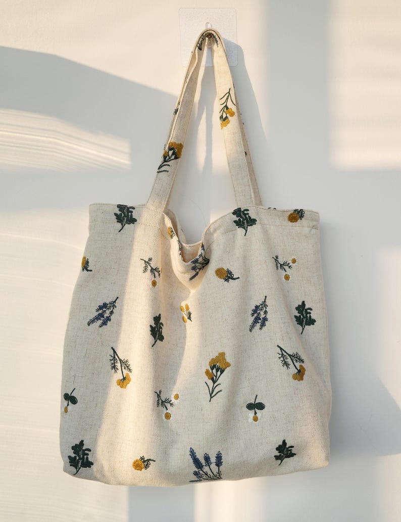 May include: Cream-colored tote bag with embroidered floral patterns in yellow, green, and blue. The bag has two long handles and is made of a textured fabric. The floral design is spread across the bag's surface.