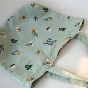 May include: A light green bonnet with a floral pattern featuring yellow, white, and purple flowers. The bonnet has matching fabric ties and is made of a soft material. The bonnet is designed for babies and toddlers.