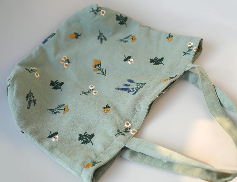 May include: A light green bonnet with a floral pattern featuring yellow, white, and purple flowers. The bonnet has matching fabric ties and is made of a soft material. The bonnet is designed for babies and toddlers.