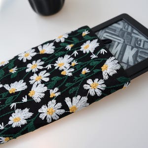 May include: Black e-reader case featuring white daisy embroidery and green stems. The case holds a black e-reader with a screen displaying text. The case is made of fabric and has a loop on the side.