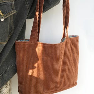 May include: A brown corduroy tote bag with long straps. The bag has a rectangular shape and a patterned interior lining. The corduroy fabric has vertical ridges, and the bag is held by a person wearing a denim jacket.