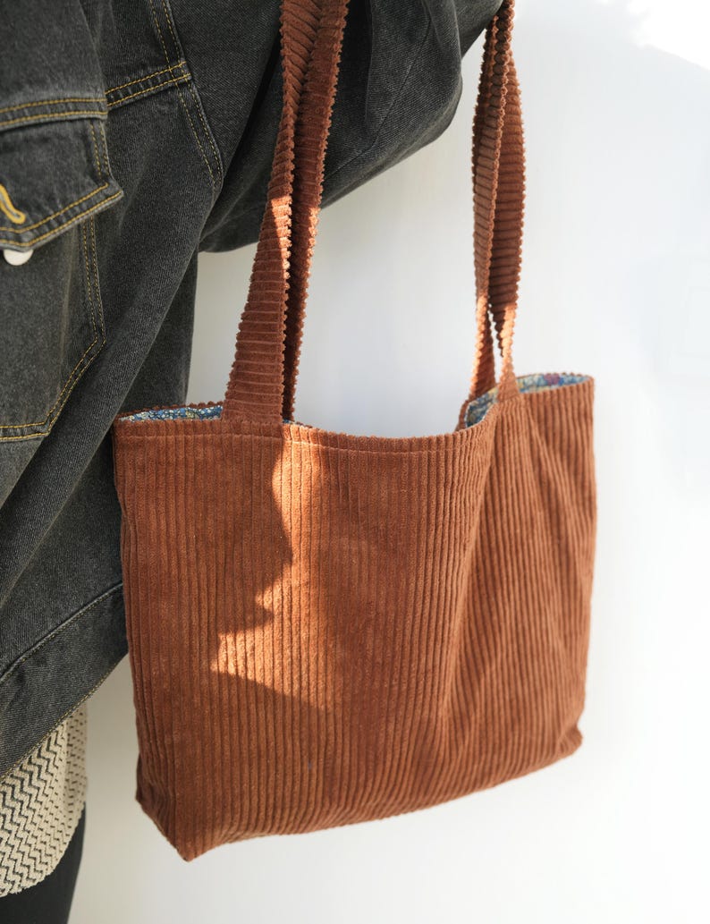 May include: A brown corduroy tote bag with long straps. The bag has a rectangular shape and a patterned interior lining. The corduroy fabric has vertical ridges, and the bag is held by a person wearing a denim jacket.