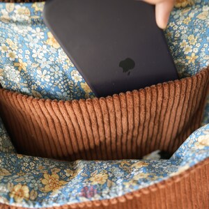 May include: Close-up of a brown corduroy pocket inside a bag. The pocket is set against a blue floral fabric with yellow and white flowers. A dark purple phone is partially inserted into the pocket, with the Apple logo visible.