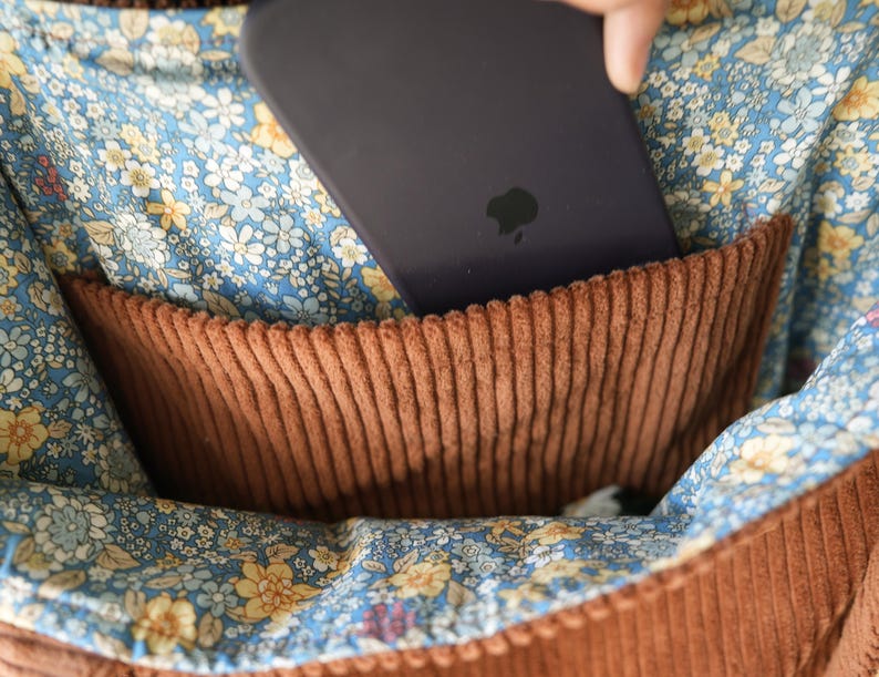 May include: Close-up of a brown corduroy pocket inside a bag. The pocket is set against a blue floral fabric with yellow and white flowers. A dark purple phone is partially inserted into the pocket, with the Apple logo visible.