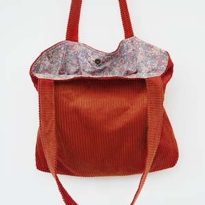 Peut inclure: Un sac fourre-tout en velours côtelé de couleur rouille avec deux bandoulières. L'intérieur est doublé d'un tissu clair orné d'un motif floral dense dans des tons de bleu, rose et vert. Un bouton-pression métallique est visible au centre.