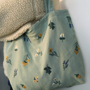 May include: A light green tote bag with embroidered floral patterns. The bag features various embroidered flowers in yellow, white, and purple. The tote bag has a simple design with a rectangular shape and two handles.
