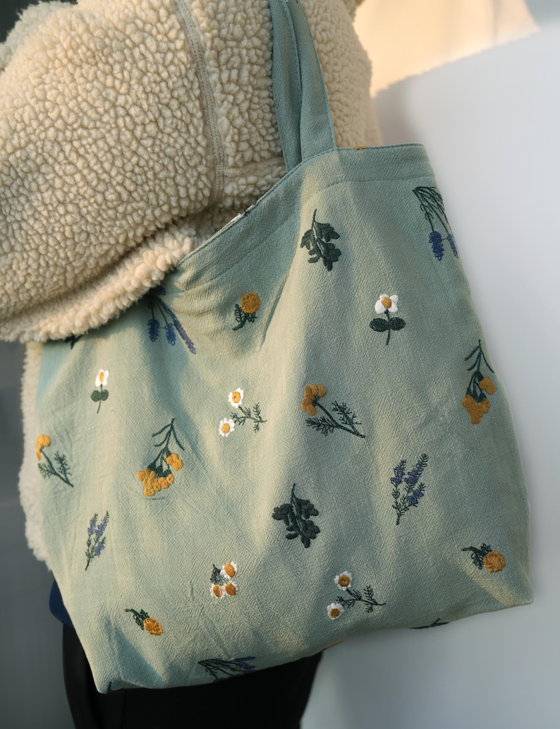 May include: A light green tote bag with embroidered floral patterns. The bag features various embroidered flowers in yellow, white, and purple. The tote bag has a simple design with a rectangular shape and two handles.