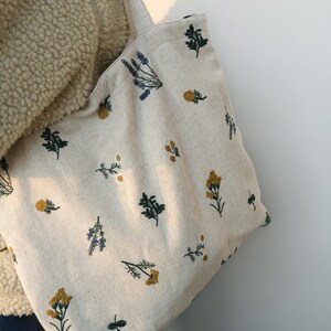 May include: Beige tote bag with embroidered floral patterns in yellow, blue, and green. The bag features a simple design with two handles and is made of a natural-looking fabric. The floral embroidery adds a touch of nature-inspired charm.