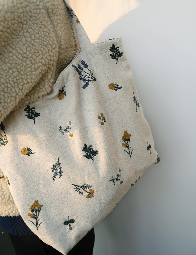 May include: Beige tote bag with embroidered floral patterns in yellow, blue, and green. The bag features a simple design with two handles and is made of a natural-looking fabric. The floral embroidery adds a touch of nature-inspired charm.