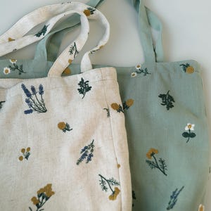 May include: Two tote bags with floral embroidery. One bag is off-white with blue, yellow, and green embroidered flowers. The other bag is light green with similar floral embroidery. Both bags have long handles.