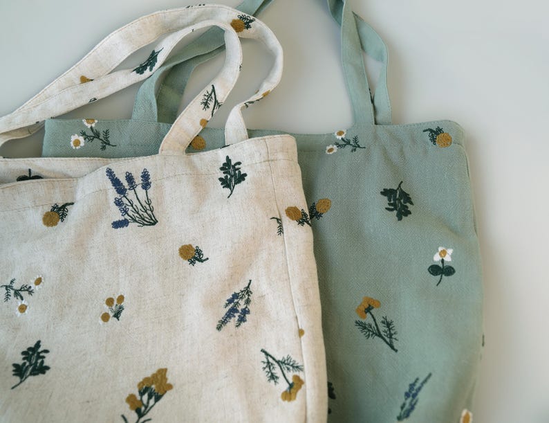 May include: Two tote bags with floral embroidery. One bag is off-white with blue, yellow, and green embroidered flowers. The other bag is light green with similar floral embroidery. Both bags have long handles.