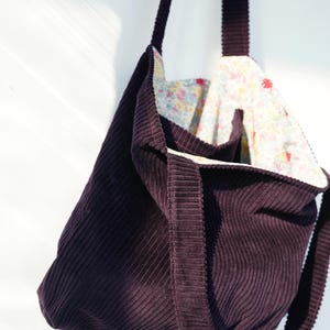 Op de afbeelding: Een donkerpaarse corduroy tote bag met een bloemenpatroon voering. De tas heeft een rechthoekige vorm met een brede band en een bijpassende corduroy buitenkant. De binnenvoering heeft een kleurrijk bloemenpatroon.