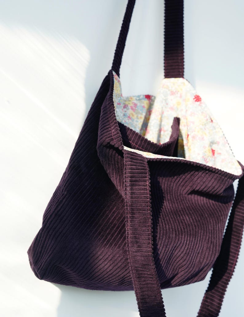 Op de afbeelding: Een donkerpaarse corduroy tote bag met een bloemenpatroon voering. De tas heeft een rechthoekige vorm met een brede band en een bijpassende corduroy buitenkant. De binnenvoering heeft een kleurrijk bloemenpatroon.