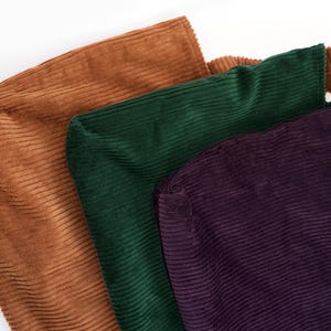 Op de afbeelding: Drie corduroy draagtassen in tinten bruin, groen en paars. De tassen zijn gemaakt van een geribbelde stof en hebben een eenvoudig, minimalistisch ontwerp. De tassen zijn waarschijnlijk bedoeld om items te dragen.