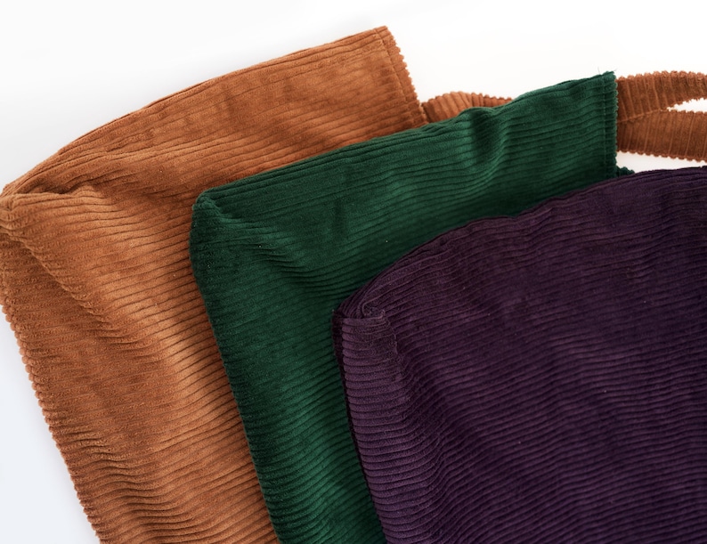 Op de afbeelding: Drie corduroy draagtassen in tinten bruin, groen en paars. De tassen zijn gemaakt van een geribbelde stof en hebben een eenvoudig, minimalistisch ontwerp. De tassen zijn waarschijnlijk bedoeld om items te dragen.