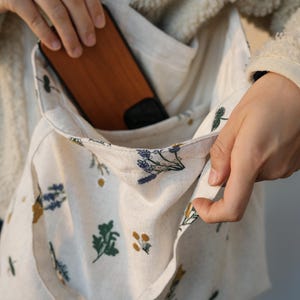 May include: Cream-colored tote bag with a floral pattern. The bag has a pocket and a wooden phone case is visible inside. The floral design includes green, yellow, and blue botanical illustrations. The bag is held by a person.